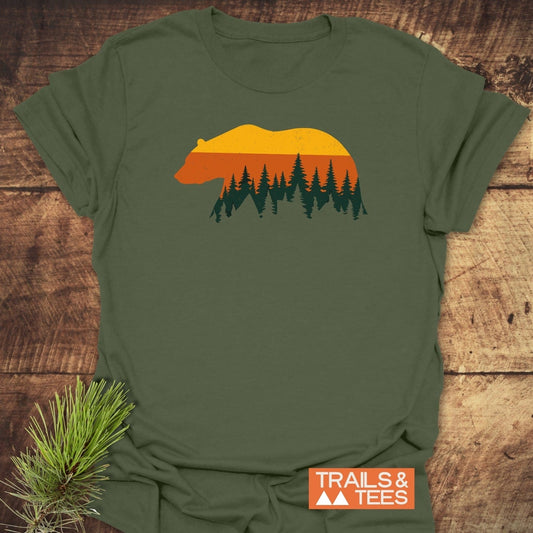 Vintage Grizzly Sunset T-Shirt featuring a bear silhouette with sunset and pine forest design, displayed flat with TRAILS & TEES logo on a wooden surface.