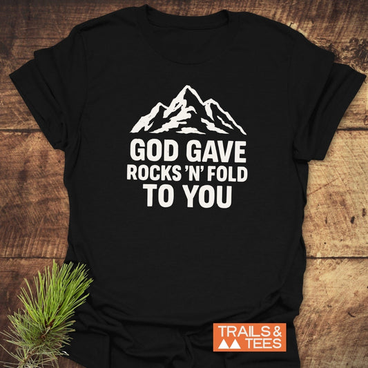 Black T-shirt with a white mountain graphic and text “GOD GAVE ROCKS ’N’ FOLD TO YOU,” featuring the Trails & Tees logo, highlighting outdoor adventure spirit.