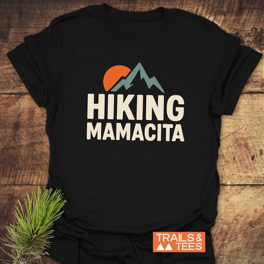 Hiking Mamacita T-Shirt featuring mountain peaks and sun graphic, laid on wood with pine sprig, showcases comfort and adventure-ready design by Trails & Tees.