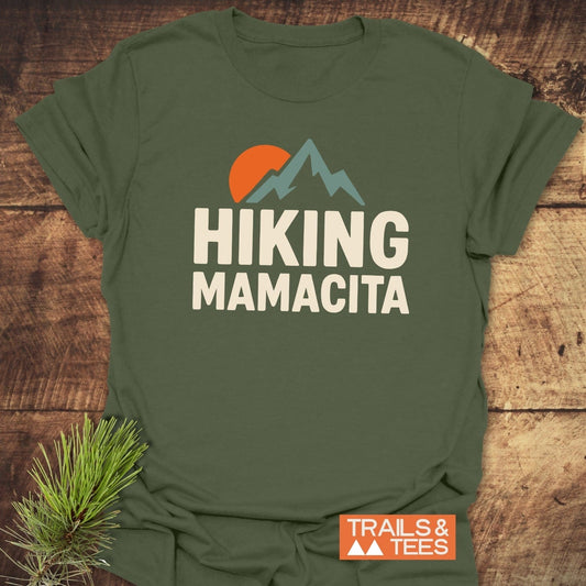 Olive-green Hiking Mamacita T-Shirt with mountain graphic and cream text, featuring TRAILS & TEES logo. Laid on wood with pine sprig.