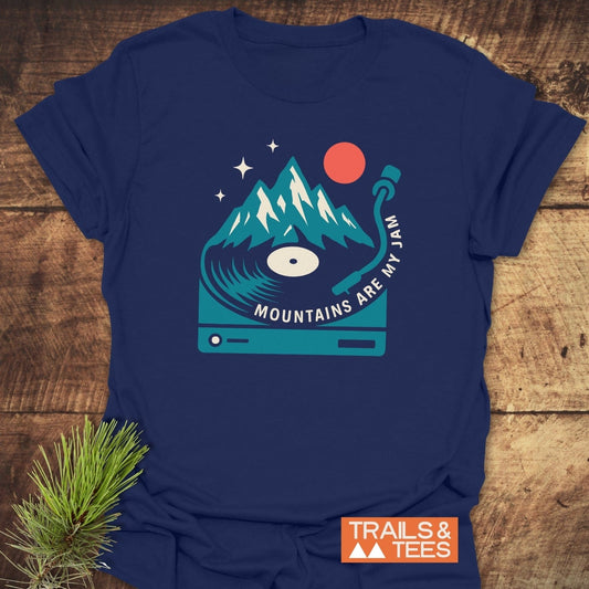 Mountains Are My Jam T-Shirt with vinyl-mountain graphic, slogan, and Trails & Tees logo, on wood surface with pine sprig, showcasing outdoor-inspired comfort and design.