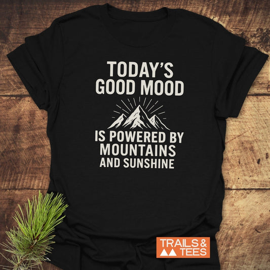 Powered By Mountains T-Shirt featuring a white mountain graphic and slogan, with an orange TRAILS & TEES. logo, laid on wood with a pine branch.