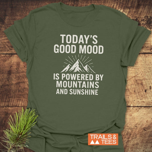Olive-green Powered By Mountains T-Shirt with mountain graphic and text, on rustic wood. Lightweight, ring-spun cotton, pre-shrunk, durable print. Trails & Tees logo visible.