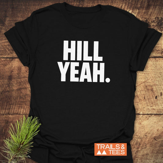 Hill Yeah T-Shirt on rustic wood, features bold white text, orange TRAILS & TEES logo, and pine twig accent. Made from soft, durable cotton.