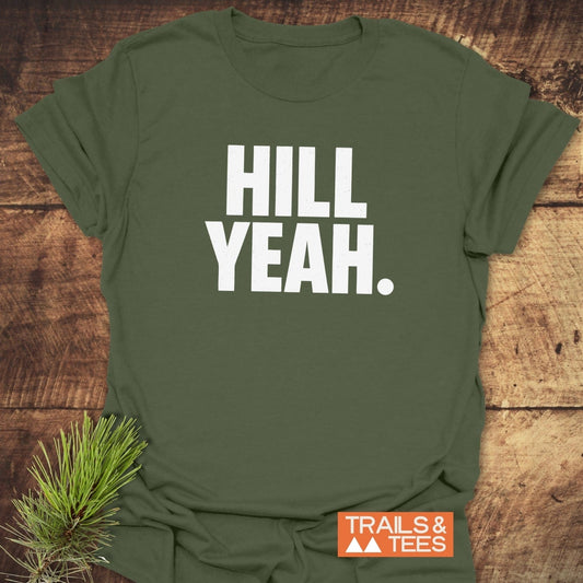 Hill Yeah T-Shirt flat-lay: Olive-green tee with HILL YEAH in white, on rustic wood. Includes TRAILS & TEES badge and pine sprig accent.