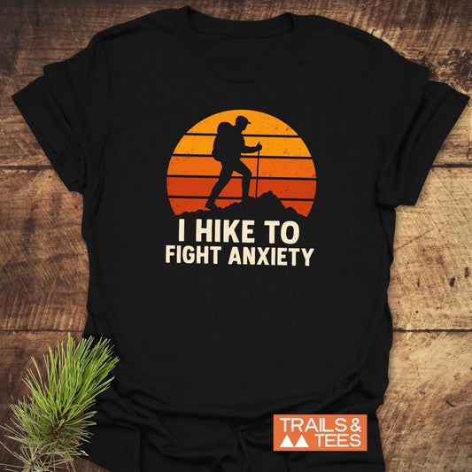 Black Hike To Fight Anxiety T-Shirt with sunset and backpacker graphic, slogan, and TRAILS & TEES branding, styled with pine sprig on wooden surface.