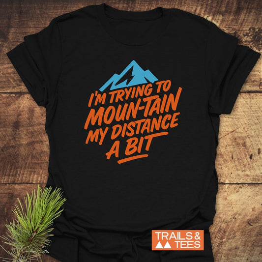 Black T-shirt featuring Moun-Tain Distance Funny T-Shirt design with blue mountain silhouette and orange text, laid on wood with pine bough, branded by Trails & Tees.