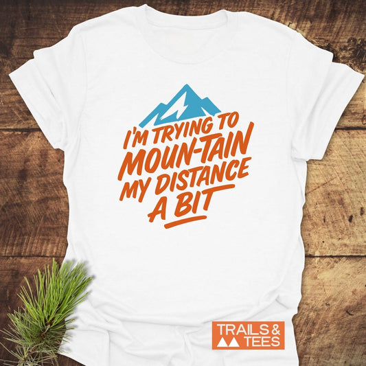 White T-shirt with blue mountain graphic and orange text: I'M TRYING TO MOUN-TAIN MY DISTANCE A BIT, by TRAILS & TEES.