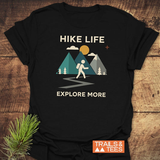 Hike Life Graphic T-Shirt with mountain scene, hiker, and EXPLORE MORE slogan, styled with a pine sprig on a wooden surface.