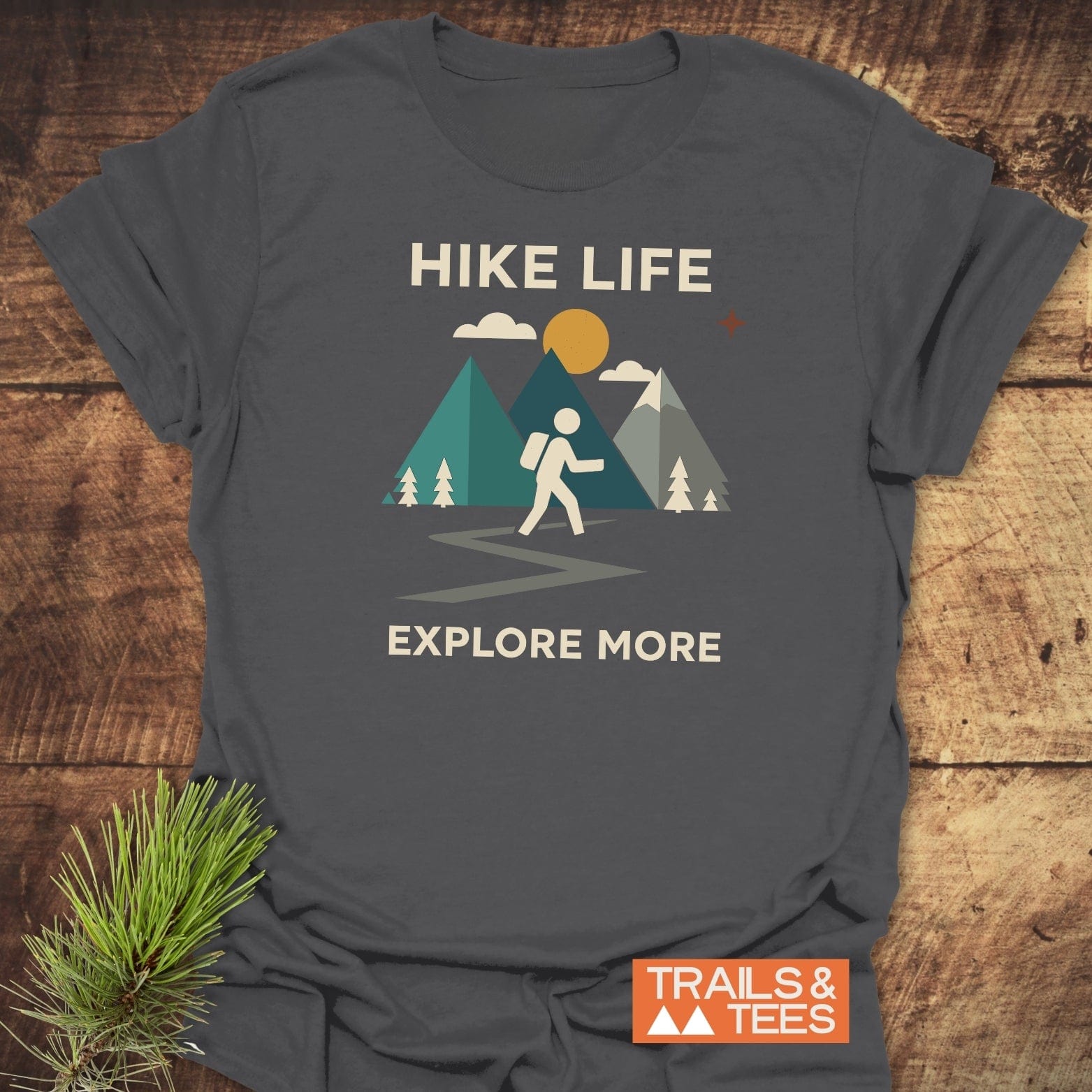 Hike Life Graphic T-Shirt featuring a stick-figure hiker, mountains, and sun graphic, with EXPLORE MORE text and TRAILS & TEES logo on a dark-gray tee.