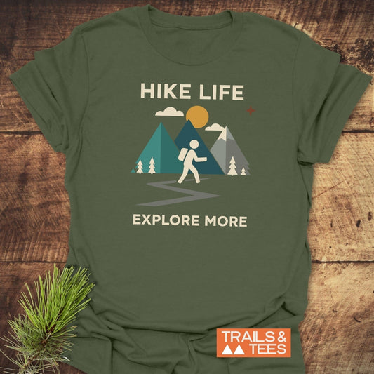 Hike Life Graphic T-Shirt featuring a hiking graphic with mountains and a sun, emphasizing comfort and adventure, laid on a wooden surface with pine sprig.