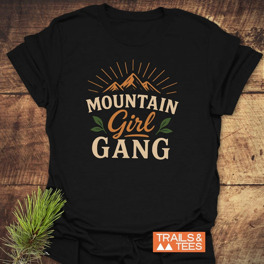 Girl Gang Hiking T-Shirt with stylized mountain graphic and “MOUNTAIN Girl GANG” text, laid on wood, featuring a rustic logo and pine sprig.