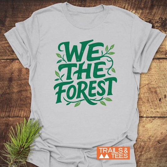 We The Forest T-Shirt with leafy green slogan on light-gray fabric, featuring a Trails & Tees logo tag, laid on a wooden surface with pine sprig detail.