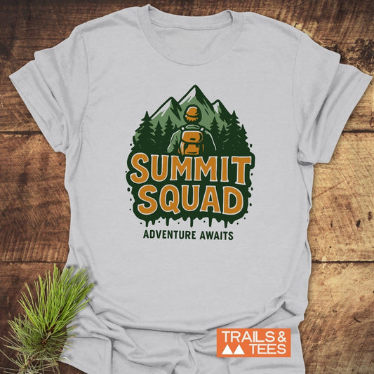Summit Squad Hiking T-Shirt featuring a hiker graphic and adventure slogan. Light cotton, DTF print, laid flat with pine sprig. Ideal for outdoor enthusiasts.