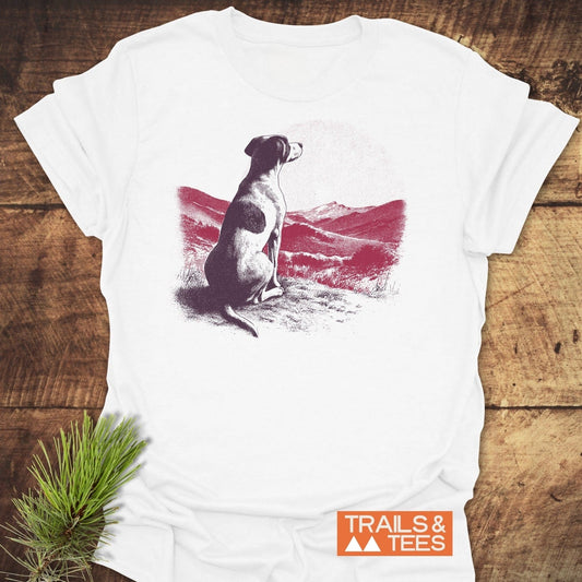 Mountains and Dog T-Shirt featuring a dog illustration with mountain backdrop, sun or moon rising, laid on wood beside pine sprig. Ideal for outdoor enthusiasts.