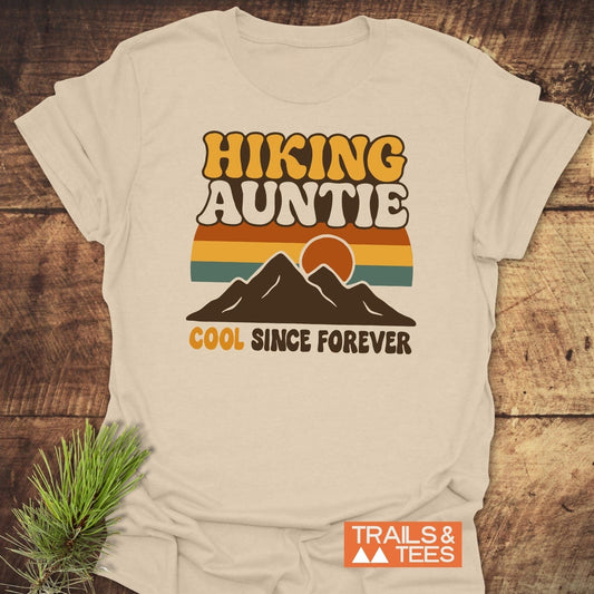 Cool Hiking Auntie T-Shirt with retro mountain graphic and slogan on soft cotton, laid on wood. Features durable print, taped shoulders, pre-shrunk fit.