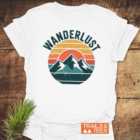 Wanderlust T-Shirt with distressed mountain graphic and logo, on wood surface with pine sprig, highlighting outdoor adventure appeal and durable, soft cotton fabric.