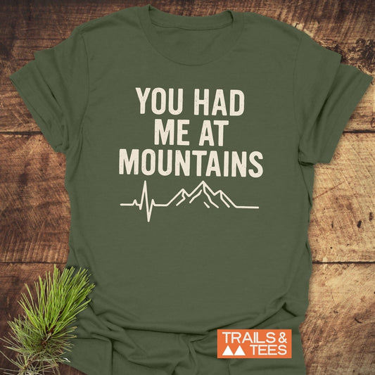 You Had Me At Mountains T-Shirt: army-green, off-white mountain graphic and text, orange “TRAILS & TEES” logo; laid on wooden surface with evergreen twig.