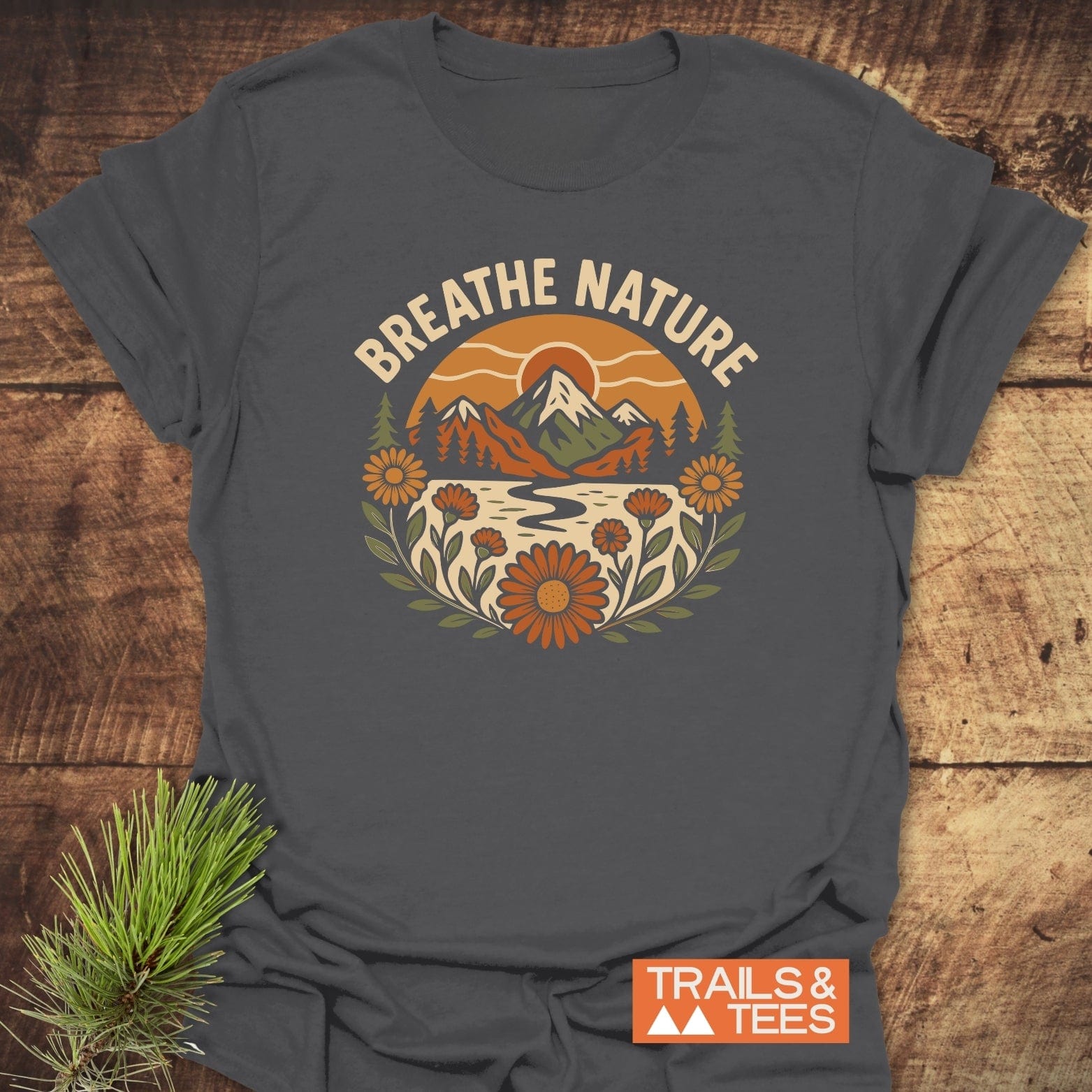 Breathe Nature T-Shirt on rustic wood, features retro nature graphic with mountains and wildflowers, showcasing a slogan. Pine sprig accentuates outdoor theme.