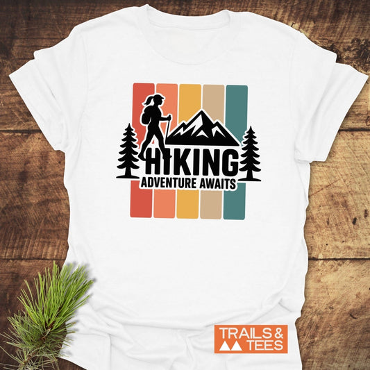 Hiking Girl T-Shirt with a mountain and hiker graphic, HIKING text, and ADVENTURE AWAITS slogan, lying on a wooden surface with pine sprig.
