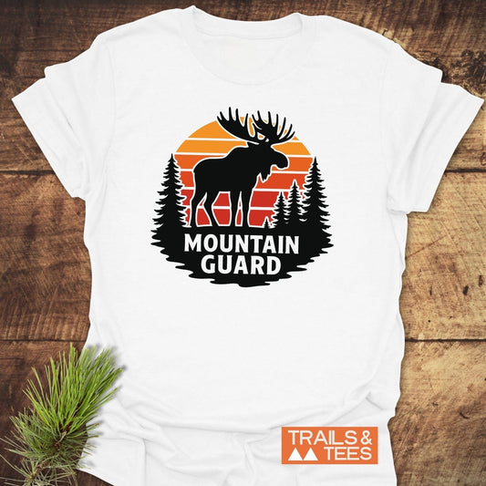 Mountain Guard Moose T-Shirt featuring a moose and pine tree graphic with a sunset backdrop, laid on wood. Includes TRAILS & TEES logo nearby.