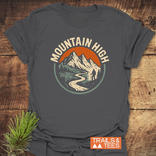 Mountain High T-Shirt with vintage mountain graphic, lone hiker silhouette, and TRAILS & TEES logo, styled with pine needles on a wooden surface.
