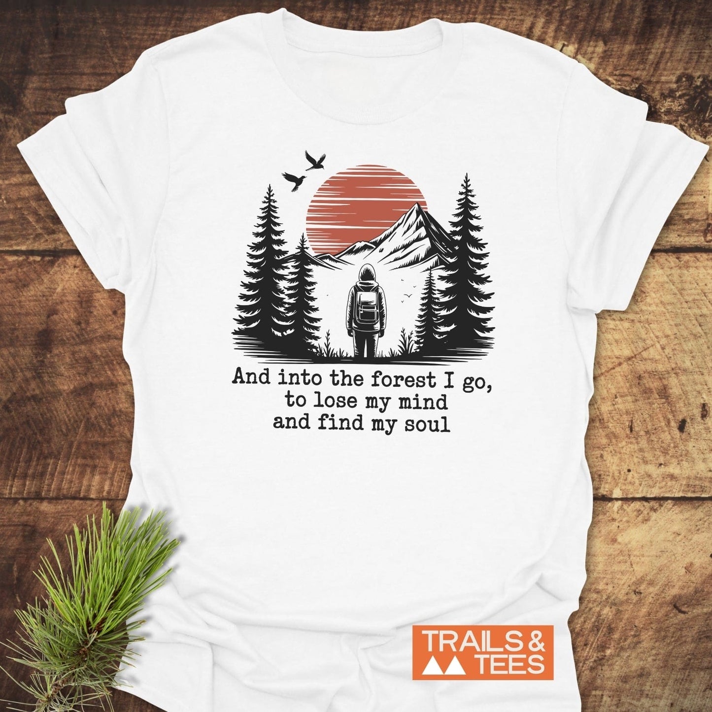 And Into The Forest I Go T-Shirt features a hiker, pine trees, mountains, and sun, with a quote beneath. Includes TRAILS & TEES. logo.