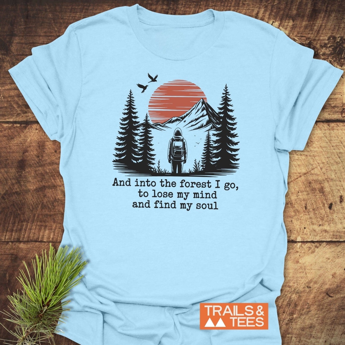 And Into The Forest I Go T-Shirt featuring a stylized outdoor scene with a backpacker, pine trees, mountains, and birds, emphasizing adventure and nature.