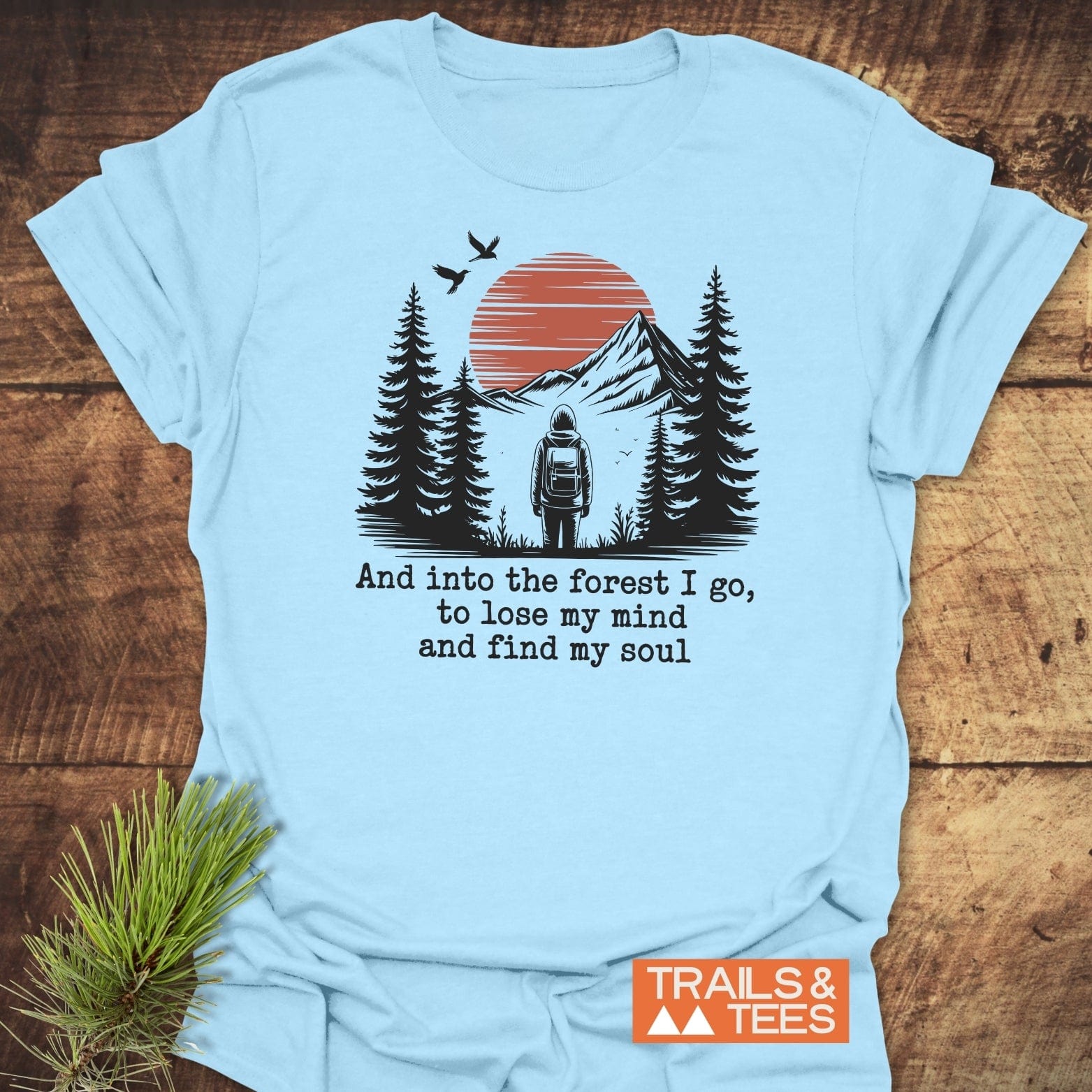 And Into The Forest I Go T-Shirt featuring a stylized outdoor scene with a backpacker, pine trees, mountains, and birds, emphasizing adventure and nature.