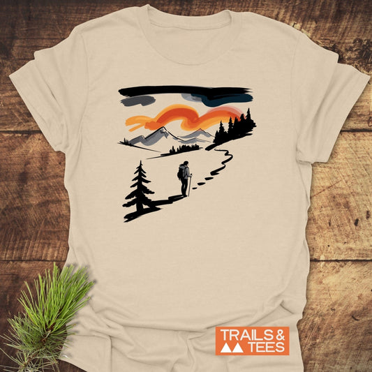 Minimalist Ink Hiking T-Shirt featuring a hiker on a trail with mountain and sunset backdrop, showcasing outdoor adventure spirit, laid flat beside a pine branch.