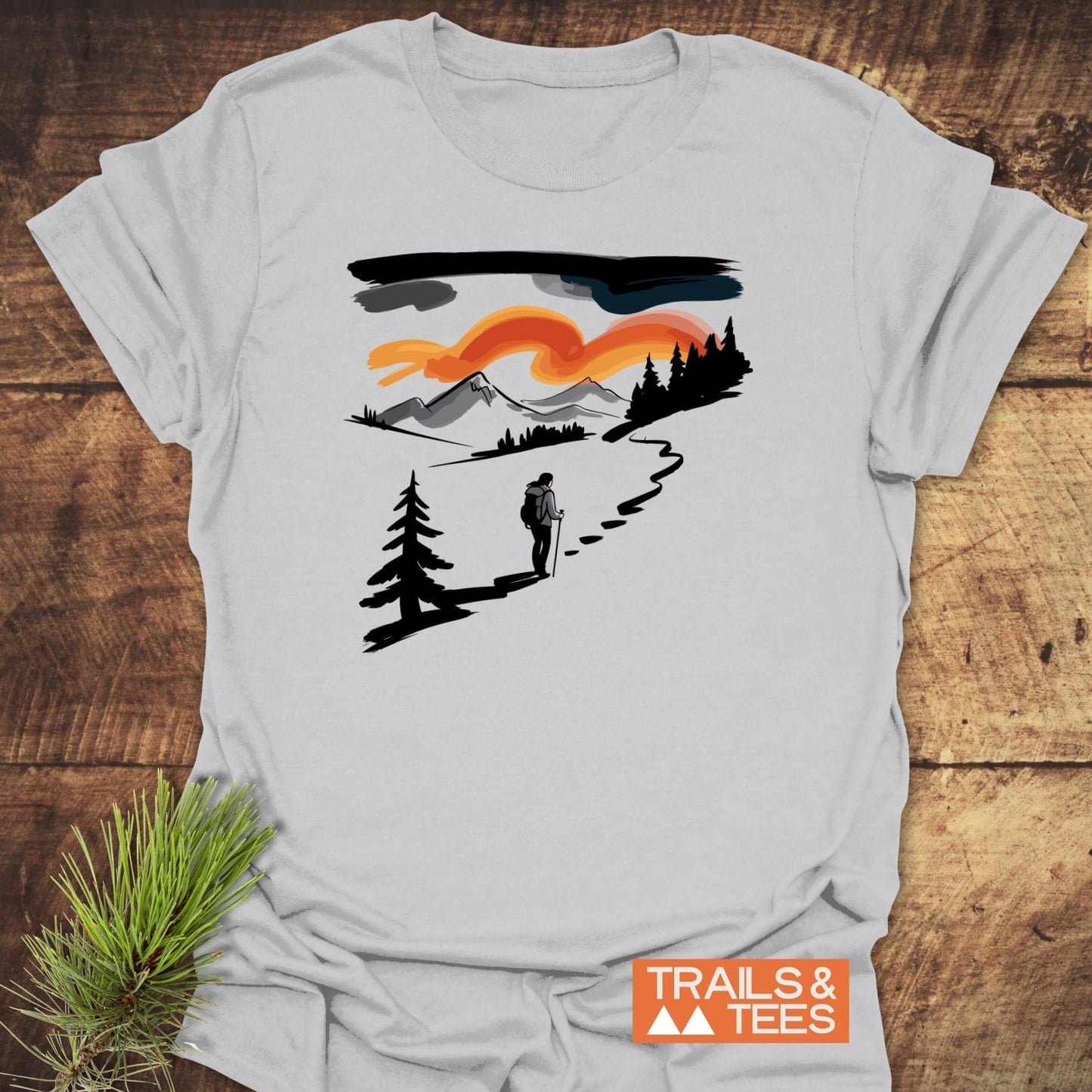 Minimalist Ink Hiking T-Shirt featuring a lone hiker graphic with mountains, pine trees, and an orange sky, laid flat with TRAILS & TEES logo visible.