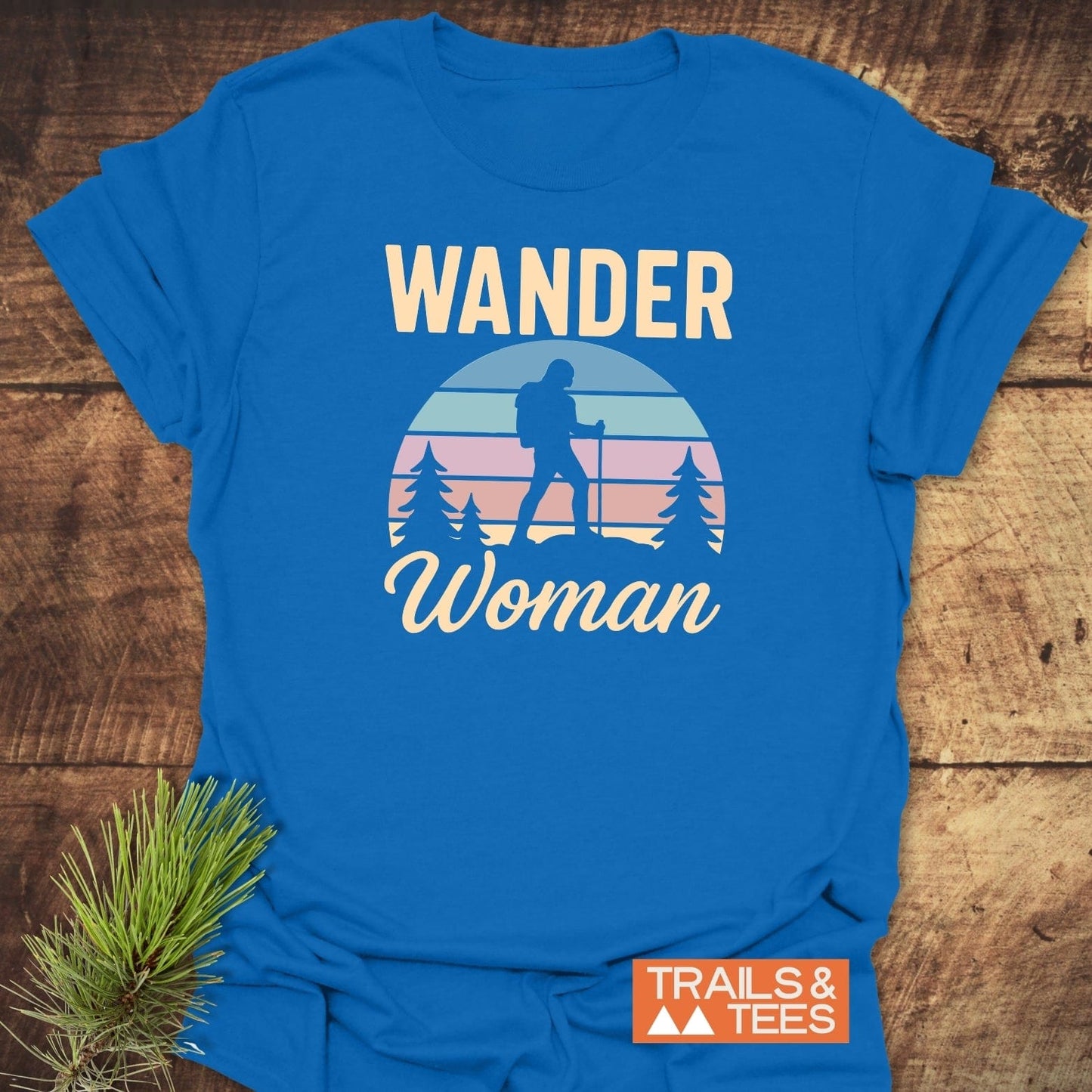 Wander Woman Retro T-Shirt featuring a hiker silhouette and pine trees against a sunset, laid on a wooden surface, showcasing outdoor-inspired design with durable prints.