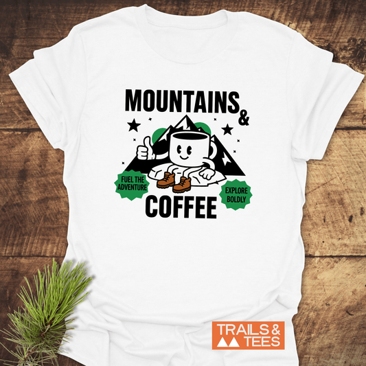 Mountains & Coffee Retro T-Shirt featuring a hiking mug graphic with mountain peaks, stars, and adventure-themed text, laid on rustic wood with pine sprig.