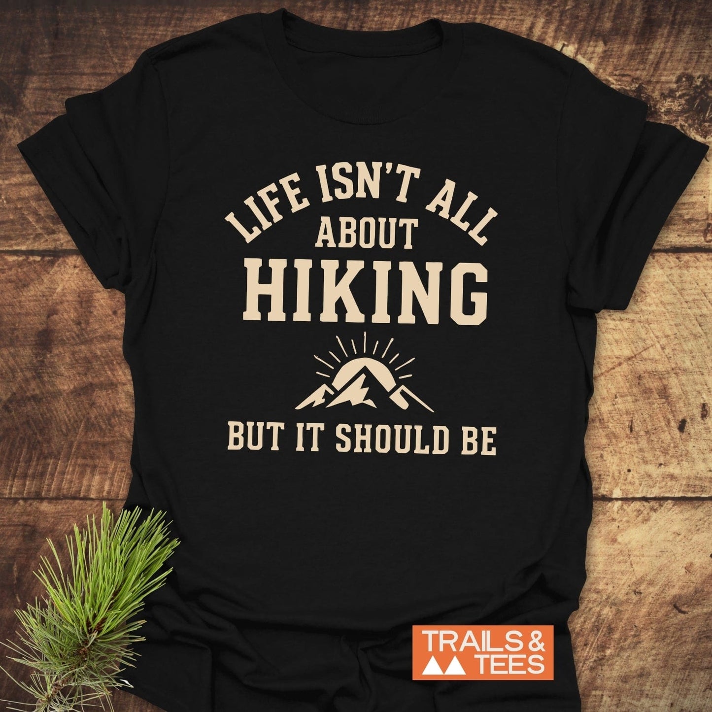 Life Isn't All About Hiking T-Shirt: Black tee with cream text and mountain graphic, orange TRAILS & TEES label, laid on wood with evergreen sprig.