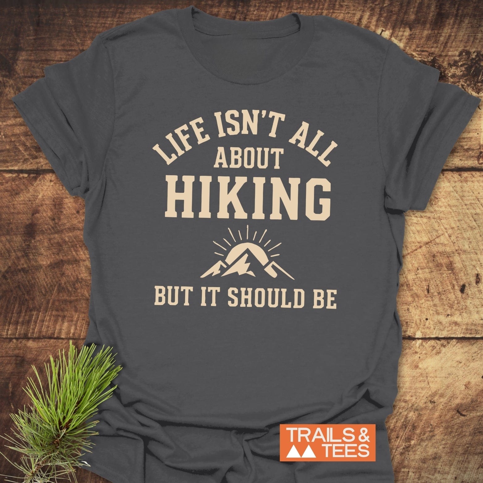 Charcoal-gray Life Isn't All About Hiking T-shirt with cream slogan and mountain icon, on wooden surface, featuring a pine sprig and TRAILS & TEES branding.