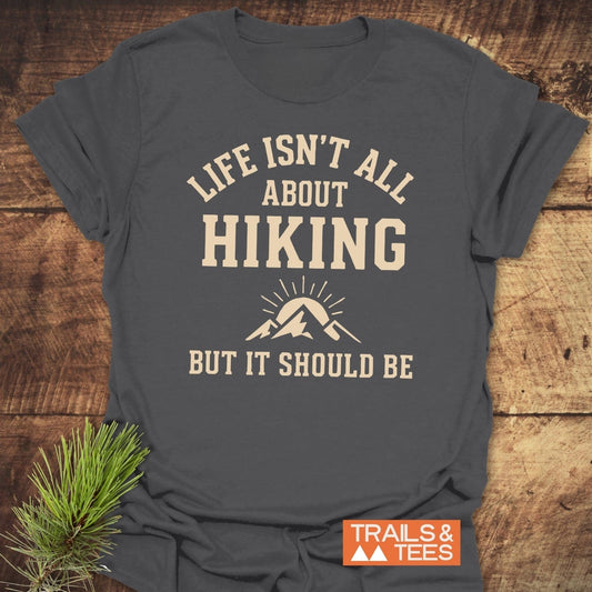Charcoal-gray Life Isn't All About Hiking T-shirt with cream slogan and mountain icon, on wooden surface, featuring a pine sprig and TRAILS & TEES branding.