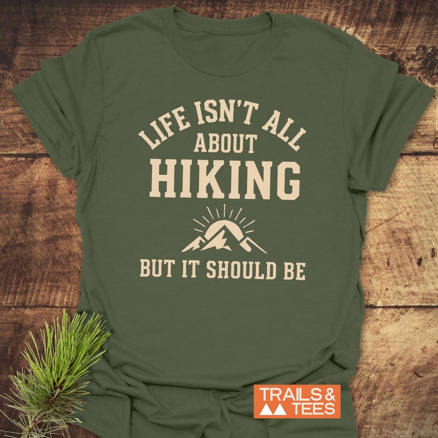 Life Isn't All About Hiking T-Shirt with mountain graphic on olive-green fabric, styled with a pine branch, promoting comfort and outdoor adventure.