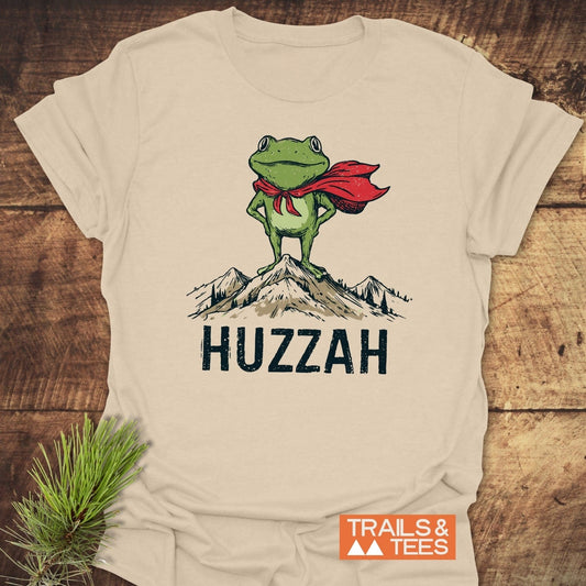 Trail Hero Funny T-Shirt features a cartoon frog in a red cape on mountain peaks with “HUZZAH” text, emphasizing adventure-ready comfort and durable design.