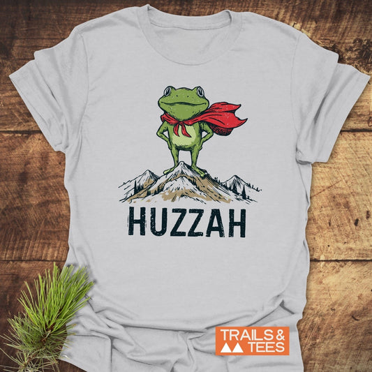 Trail Hero Funny T-Shirt featuring a cartoon green frog in a red cape atop a mountain, with “HUZZAH” text, showcasing adventure-ready design and comfort.