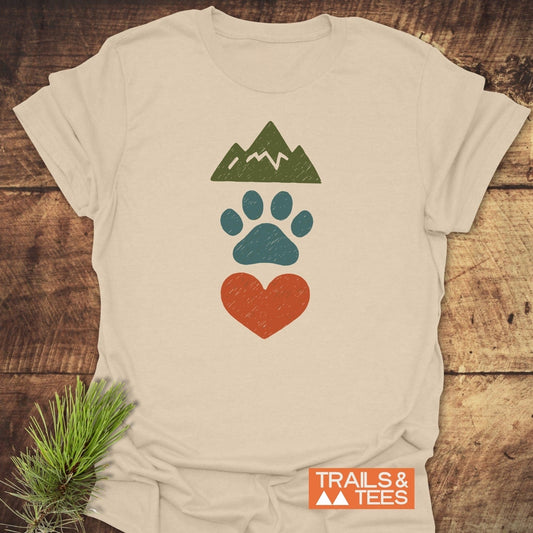 Mountains & Dogs Love T-Shirt on wood, featuring mountain, paw print, and heart graphics. Made of soft, lightweight cotton for outdoor adventures. Brand: TRAILS & TEES.
