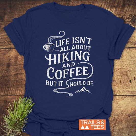 Navy-blue Hiking and Coffee T-Shirt with white slogan, coffee cup, and mountain design. Orange TRAILS & TEES logo on rough wooden background.