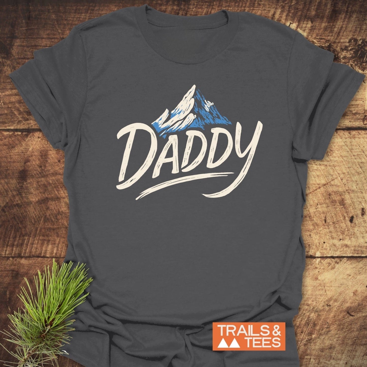 Mountain Daddy T-Shirt featuring a mountain graphic and DADDY text on soft, charcoal-gray cotton, displayed on wood with a pine branch, Trails & Tees logo visible.