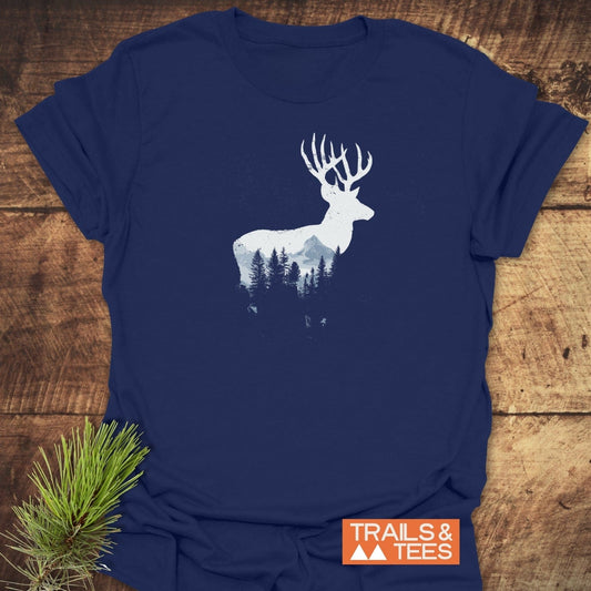 Deer Silhouette T-Shirt featuring a white buck design with mountain and pine-tree details on navy-blue cotton, laid on wood with TRAILS & TEES logo.