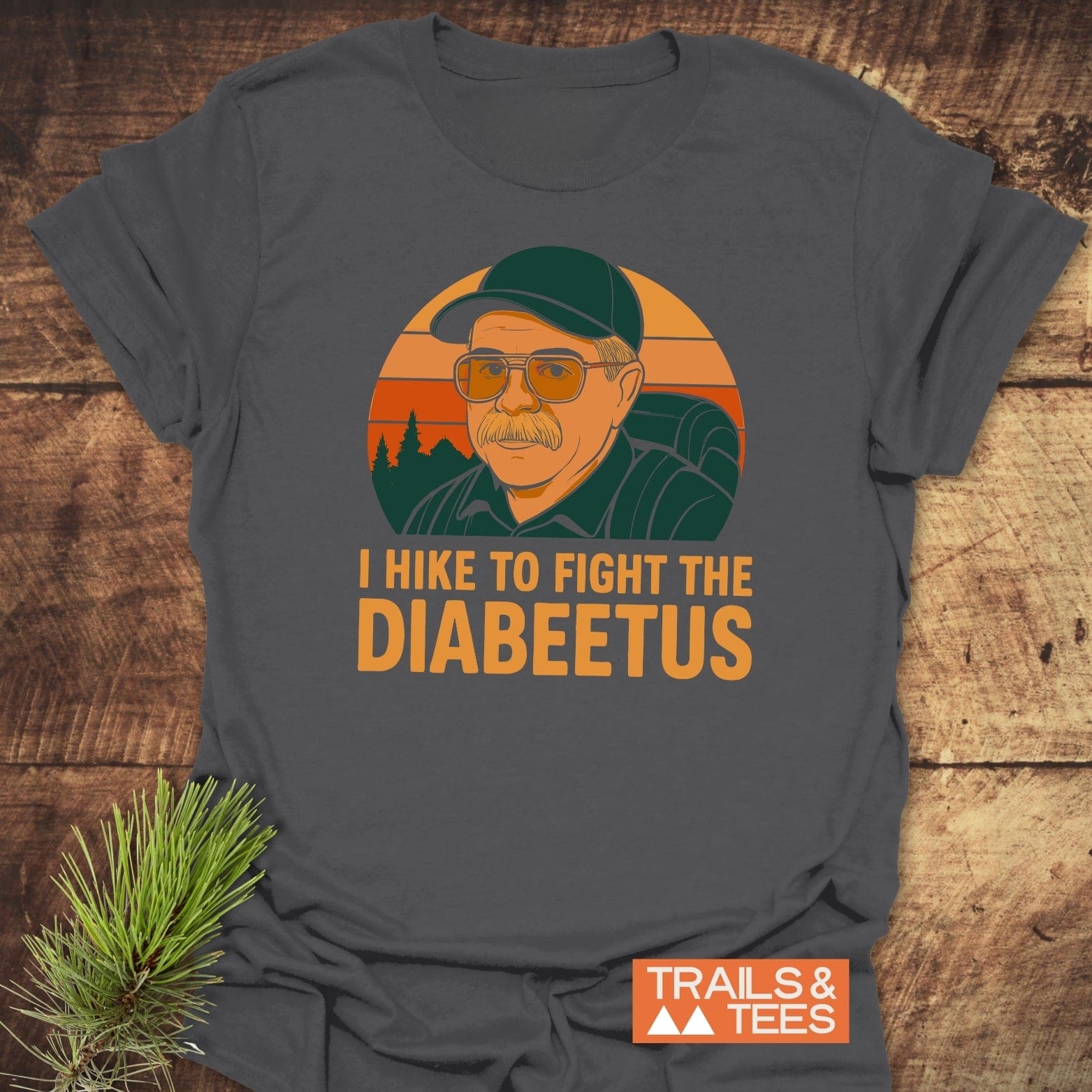 Retro Diabeetus Funny T-Shirt featuring a sunset forest scene with a hiker graphic and slogan, laid on wood with pine branch, emphasizing adventure and comfort.
