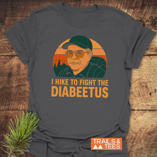Retro Diabeetus Funny T-Shirt featuring a sunset forest scene with a hiker graphic and slogan, laid on wood with pine branch, emphasizing adventure and comfort.