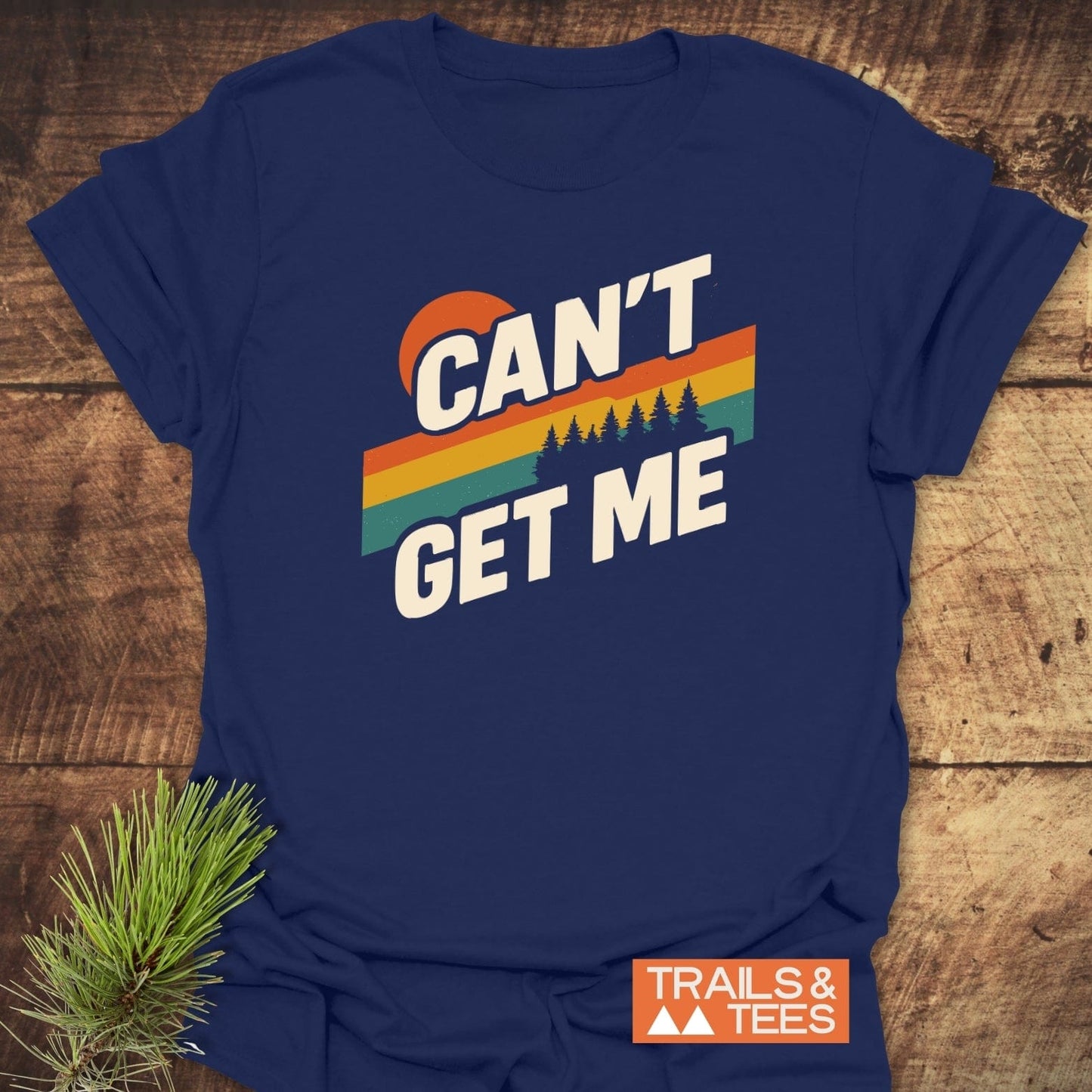 Can't Get Me Hiking T-Shirt featuring a retro graphic with sun, stripes, and trees, laid on wood. Includes “TRAILS & TEES” badge and pine branch.