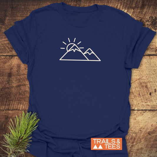 Minimalist Mountains And Sun T-Shirt on rustic wood, featuring white mountain peaks graphic. Includes TRAILS & TEES logo, highlighting comfort and adventure-ready design.