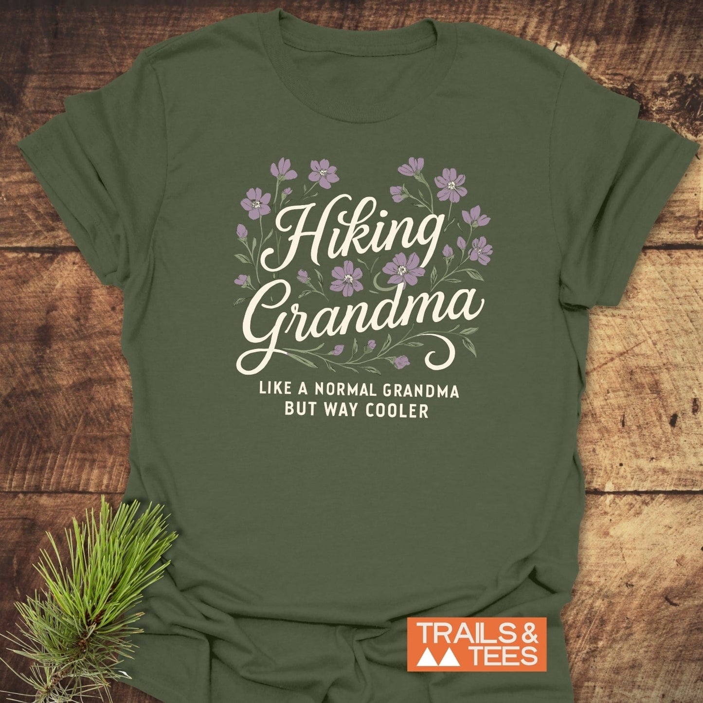 Olive-green Hiking Grandma Floral T-Shirt, featuring a purple blossom motif and cream script, styled with a pine sprig; crafted for comfort and adventure.