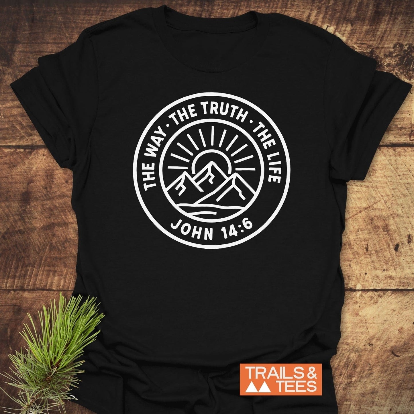 The Way, The Truth, The Life T-Shirt featuring a mountain graphic and biblical quote, laid on wood with a pine sprig, by TRAILS & TEES.