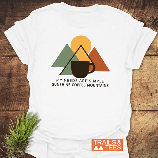 The Simple Joys T-Shirt on wood, featuring mountain-inspired triangles, sun, coffee cup graphic, and caption. Ideal for outdoor enthusiasts, crafted for comfort and durability.